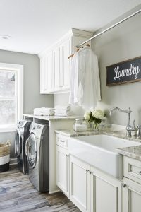 A white farmhouse apron sink gives this laundry room a classic touch, while creamy white cabinets contrast beautifully against wood grain-inspired floor tiles.