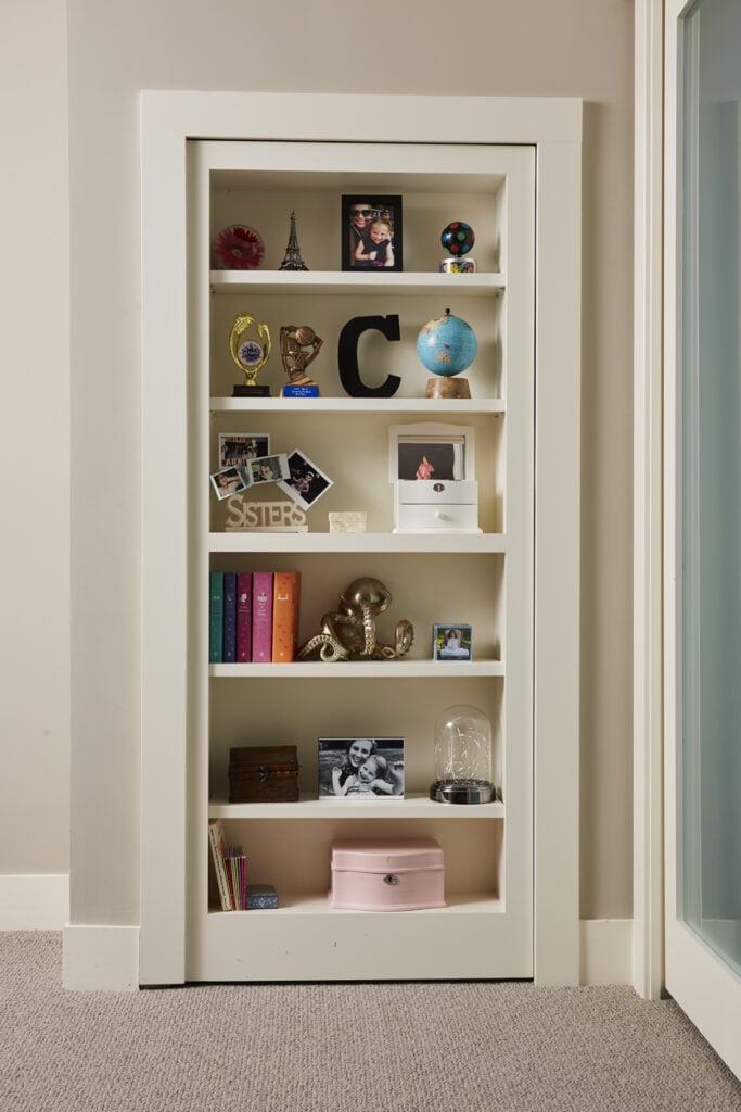 A hidden door disguised as a bookcase provides space to display beloved items and photos, adding a unique and playful decor touch to the room. (Interior Design: Carla Bast | Sneak Peek Design)